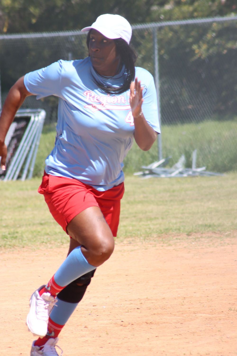 Female athlete in red shorts and white cap sprinting on a dirt field during an outdoor softball game.