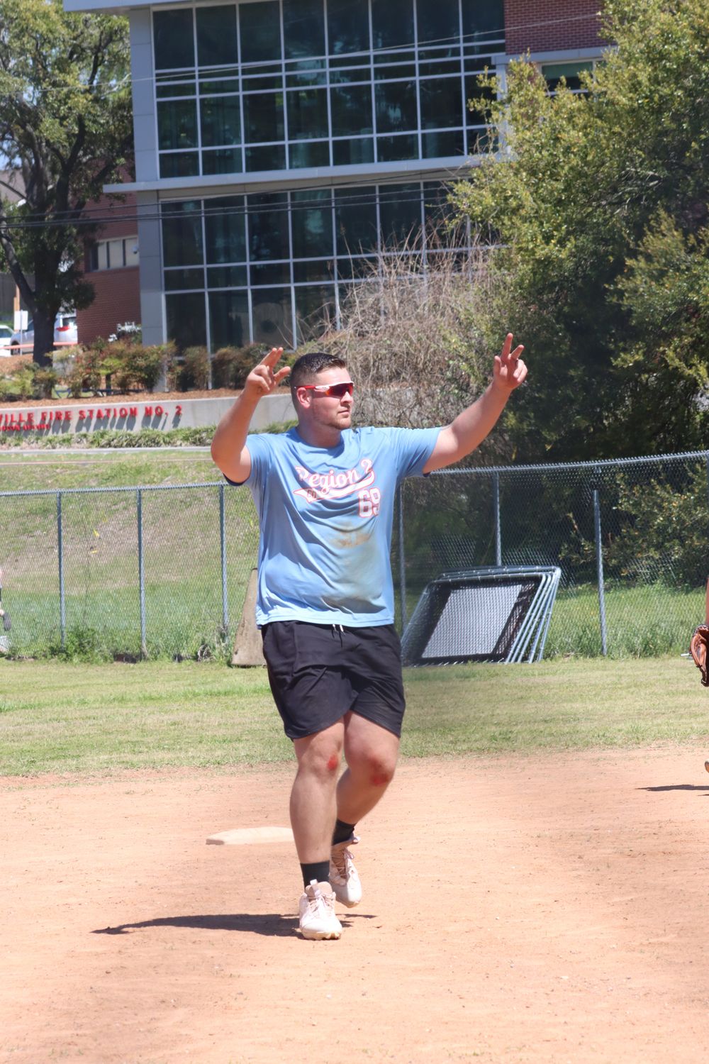 Man in light blue t-shirt and sunglasses celebrates on baseball field near base, arms raised, with building in background.