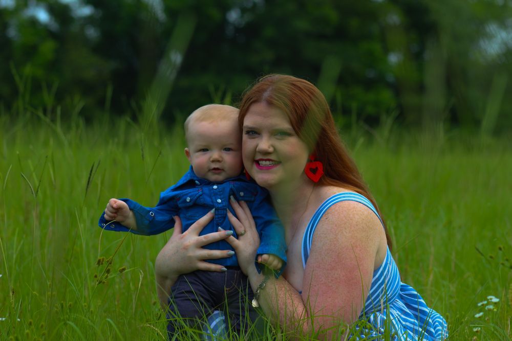 A person in a blue top sits in a green meadow on a sunny day, enjoying the outdoors with a small child.