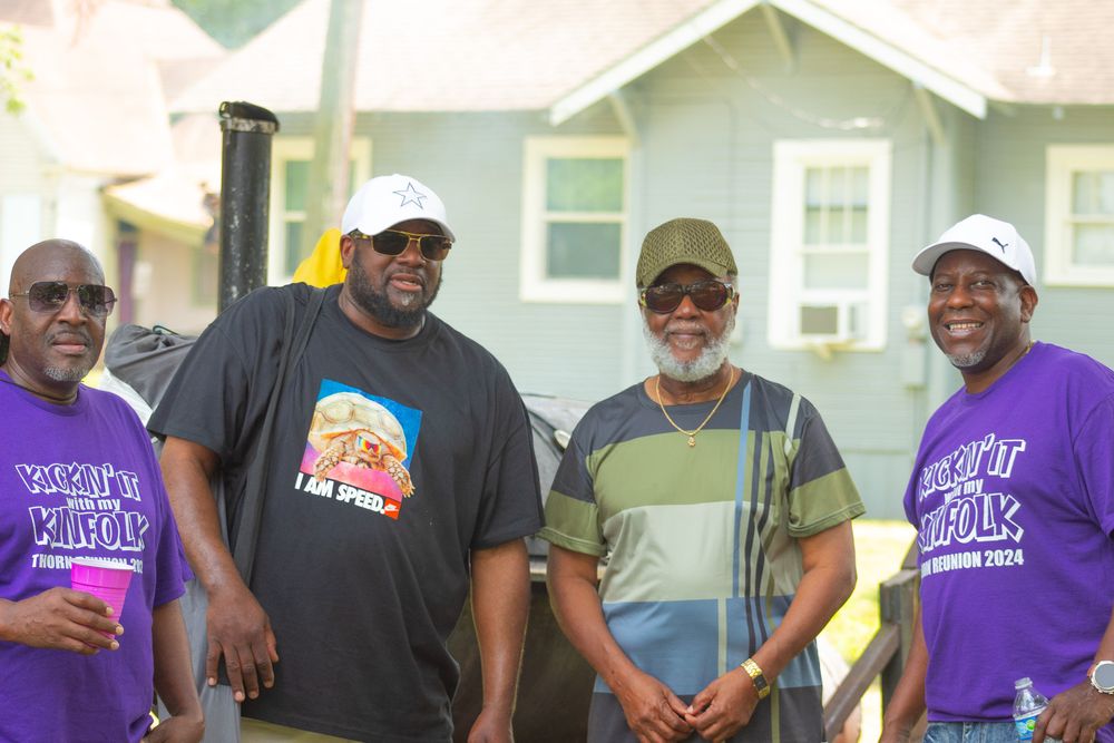 Four men wearing casual t-shirts and hats stand together smiling outdoors in front of a house.