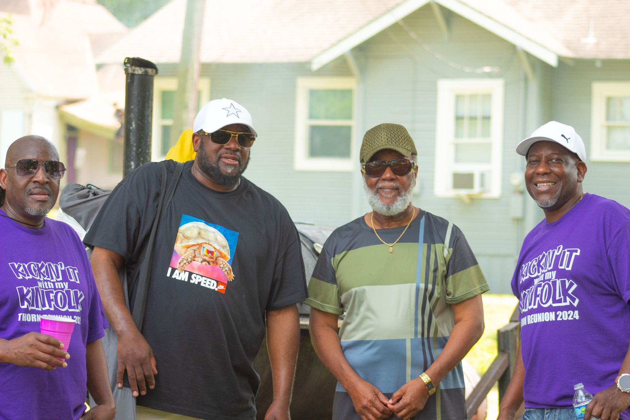 Four men wearing casual t-shirts and hats stand together smiling outdoors in front of a house.