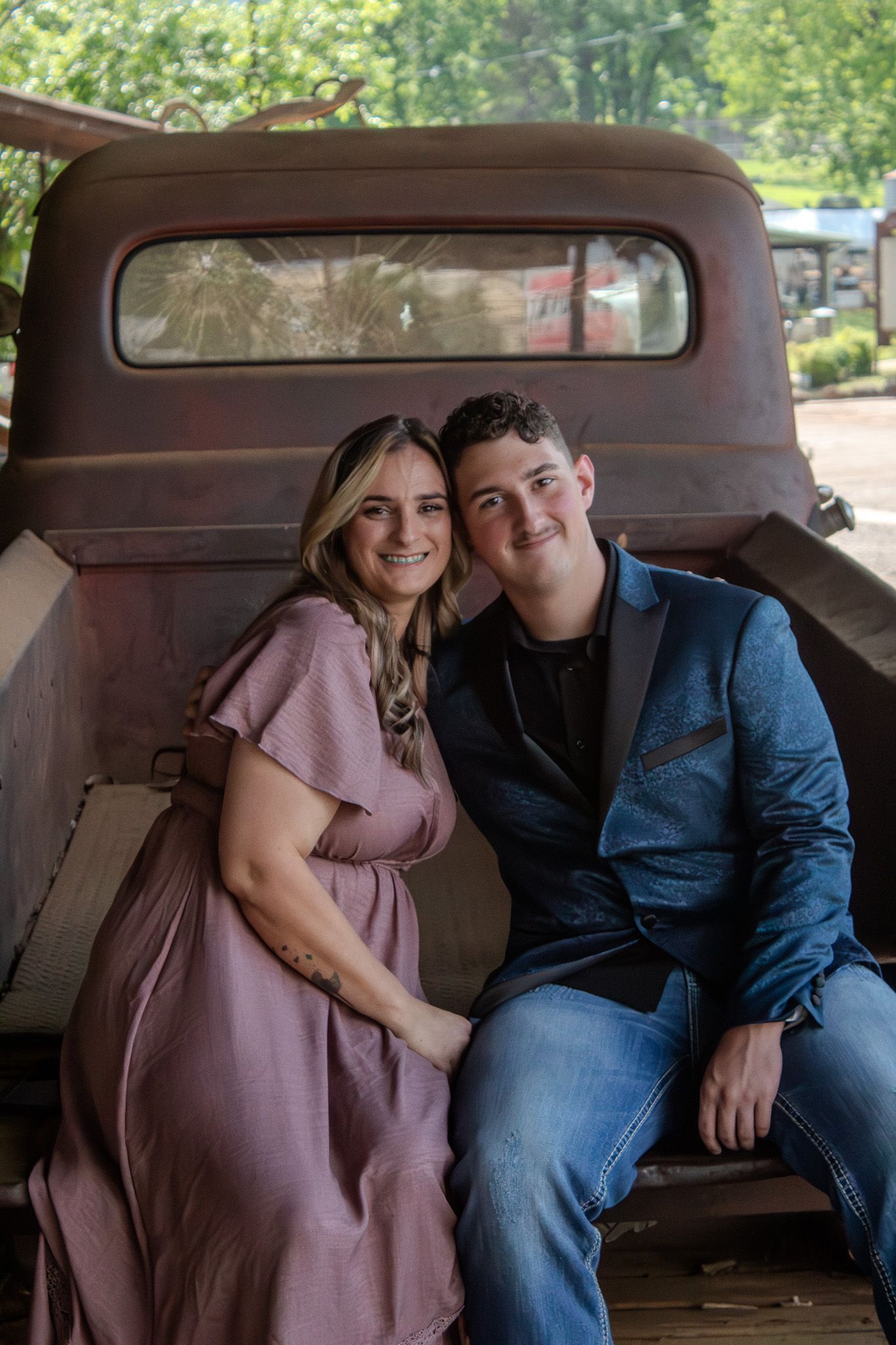 A couple in casual attire poses together inside a vintage car, with the person in pink leaning against their partner.