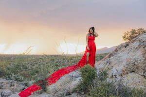 Long red dress creates striking silhouette against sunset desert mountains and saguaro cacti.