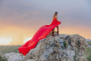 Red dress billows dramatically on desert hilltop with saguaro cacti at sunset.