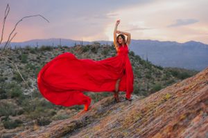A flowing red dress billows dramatically against a desert landscape at sunset with mountains in the background.