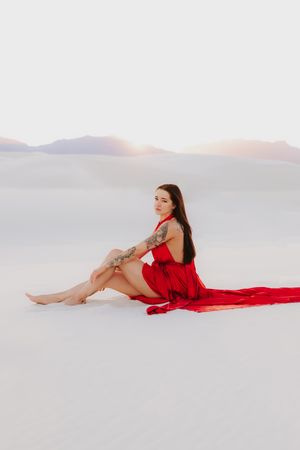 Red dress poses dramatically on white sand dunes with mountains silhouetted in the background.