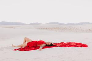 A figure in a flowing red dress poses gracefully on white sand dunes at sunset.