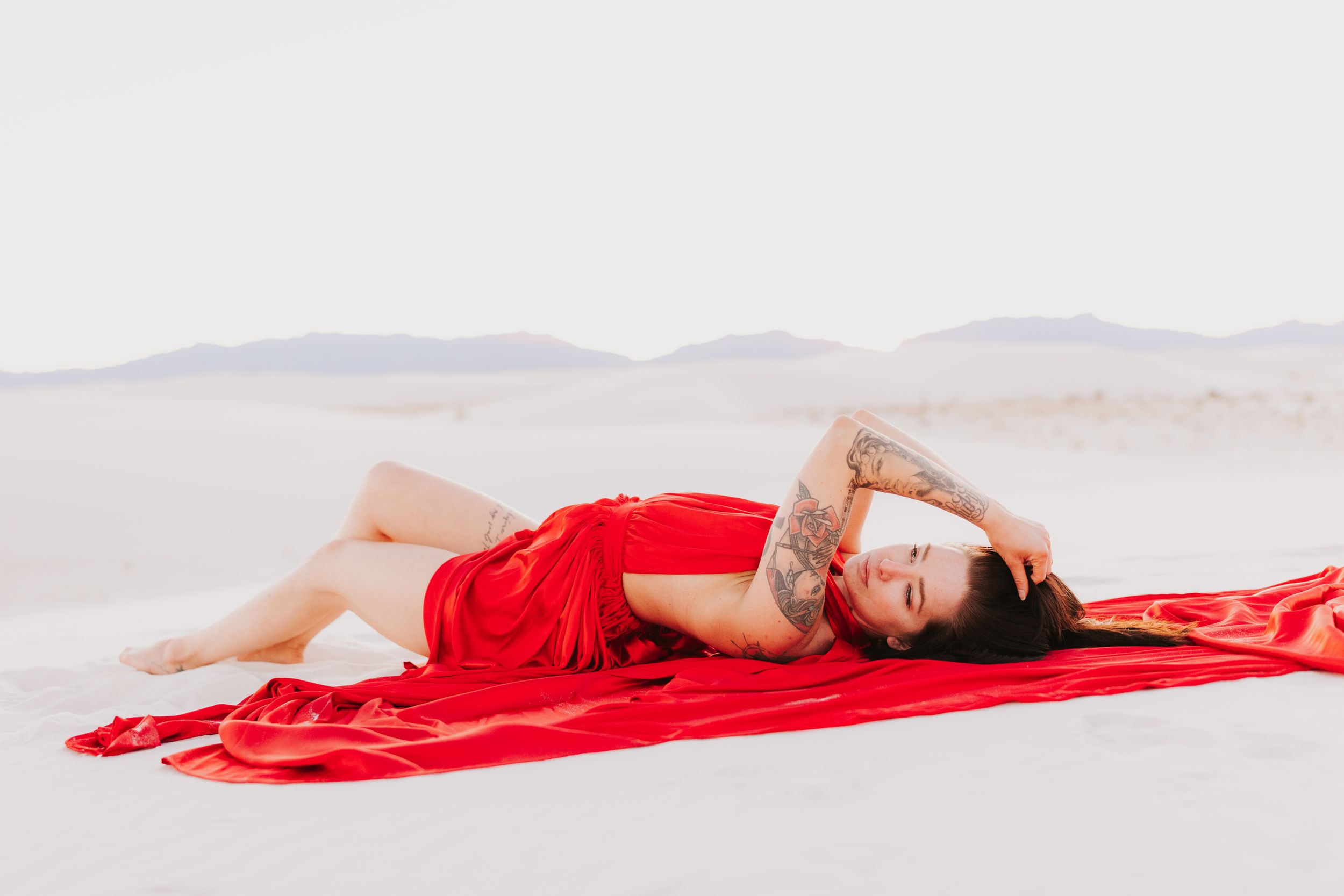 A figure in red fabric lies artistically across pristine white sand with mountains in the distance.