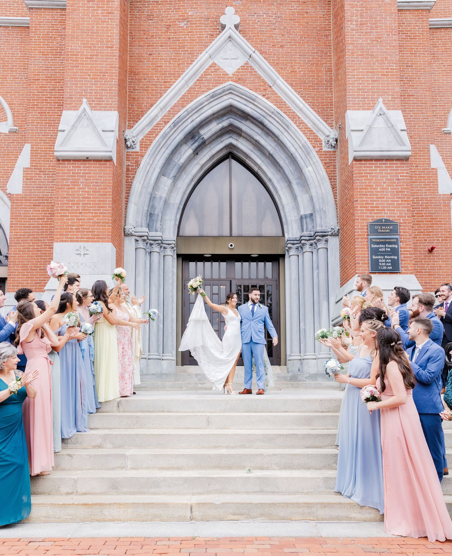 Wedding attendees in pastel dresses line church steps while flower petals are tossed in celebration.