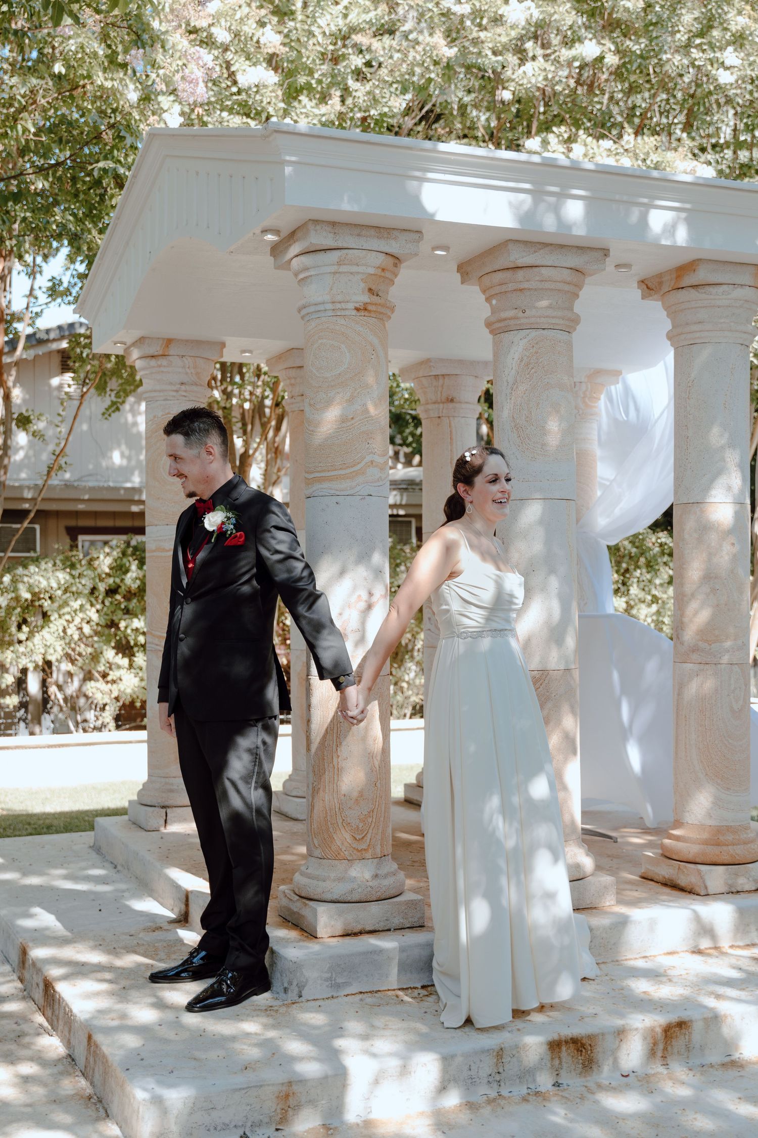 A couple poses between white columns in an outdoor garden setting.