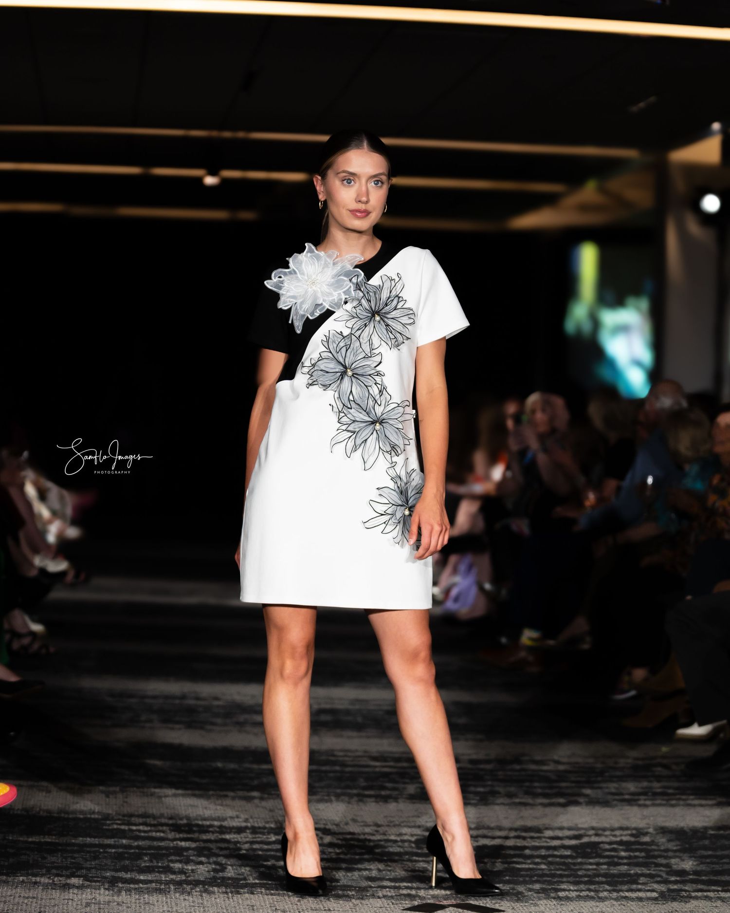 Model walks runway in white mini dress with black floral design detail at fashion show event.