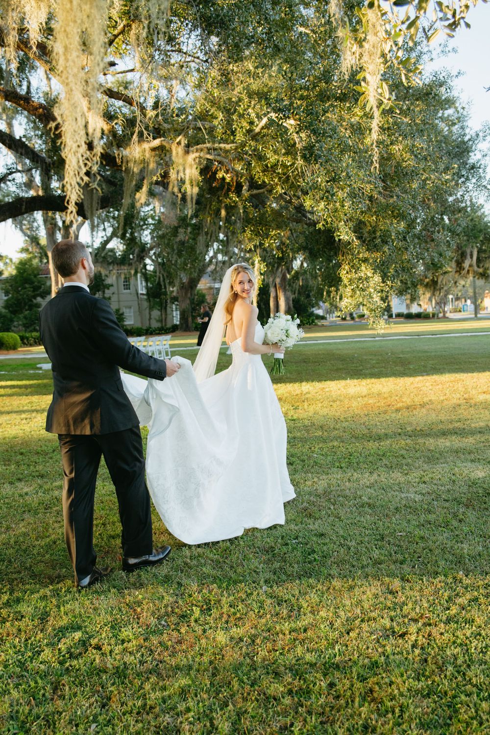 Newlyweds share a romantic moment under moss-draped trees on a sunny lawn.
