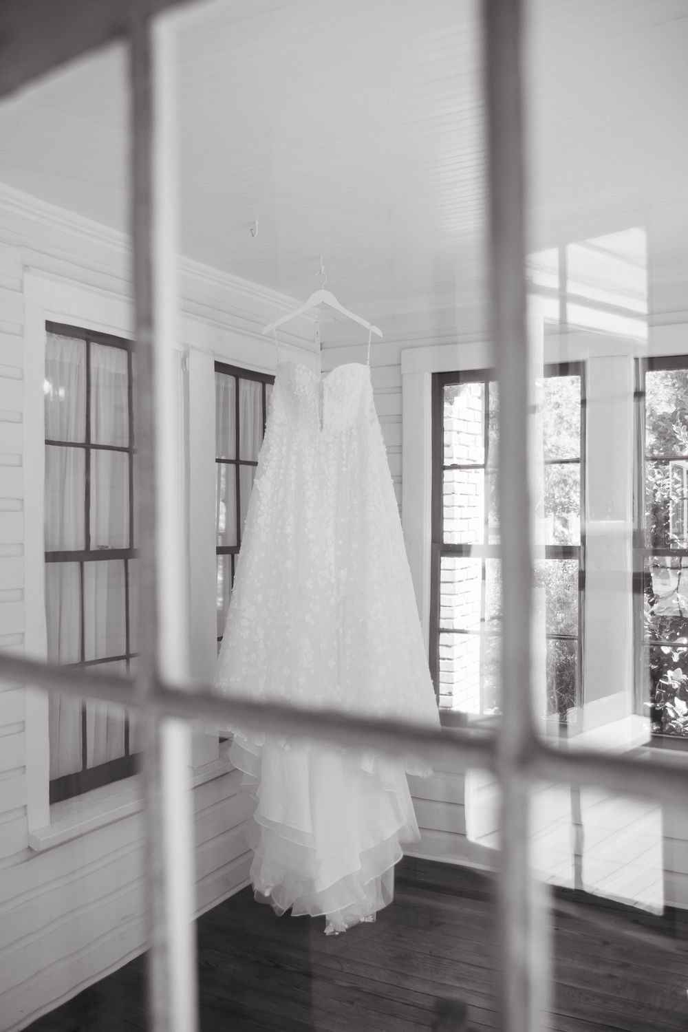 Wedding dress hangs in white room with natural light streaming through windows.