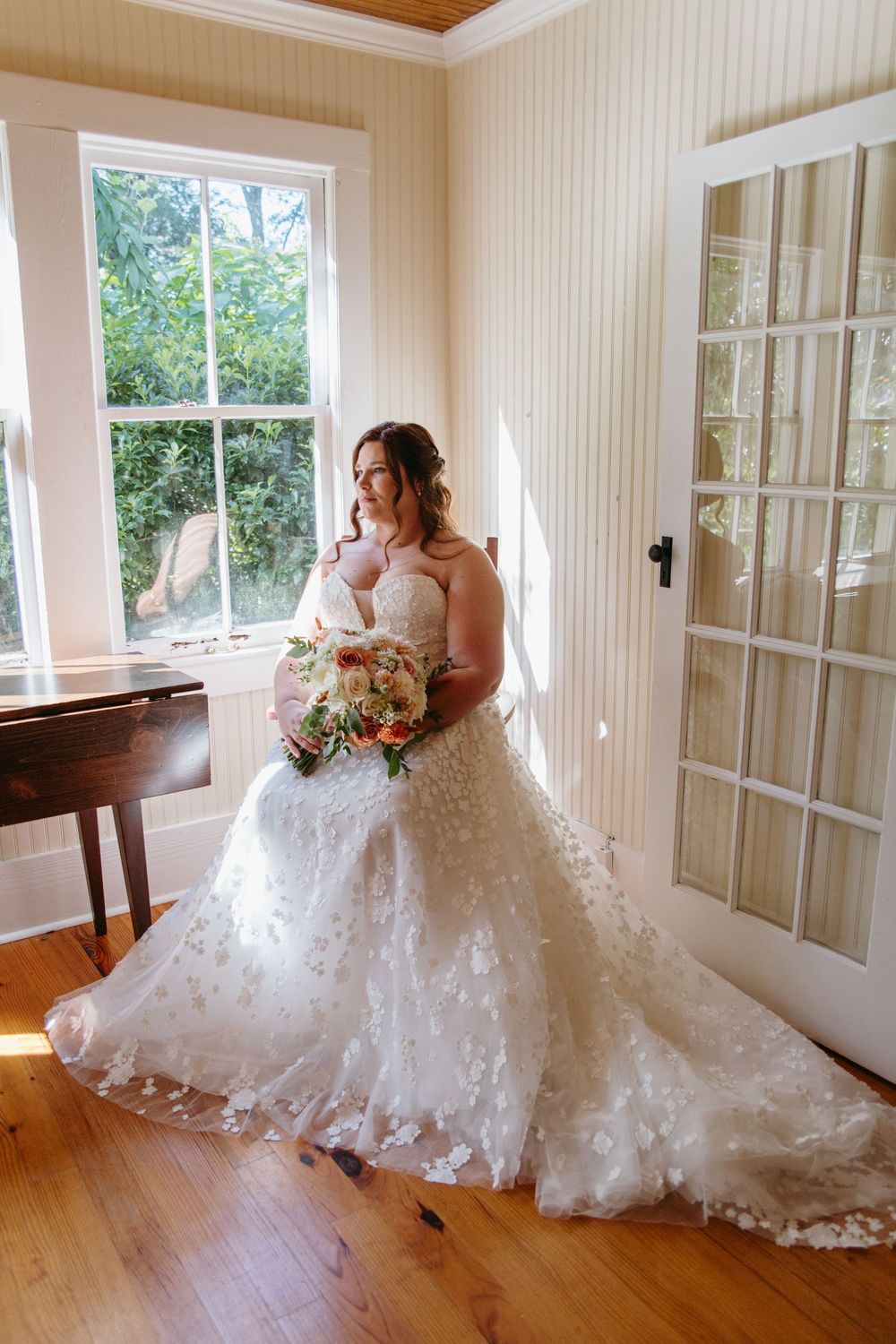 Bride in flowing lace gown poses in sunlit room with hardwood floors.