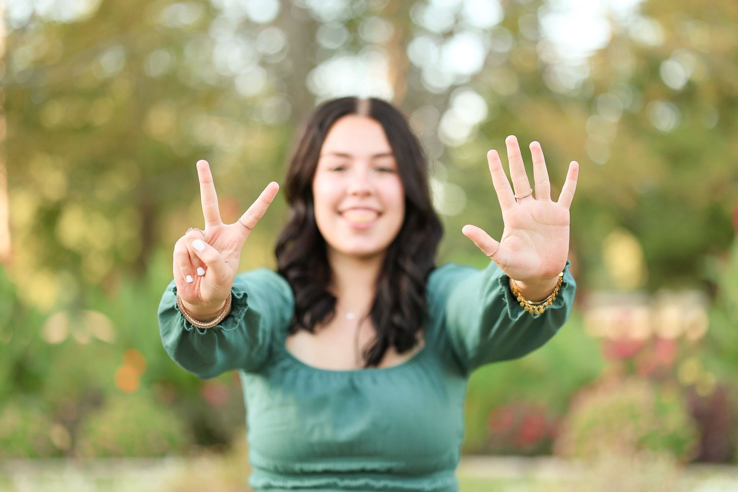 A person in a green sweater makes peace signs and smiles outdoors in a park setting.