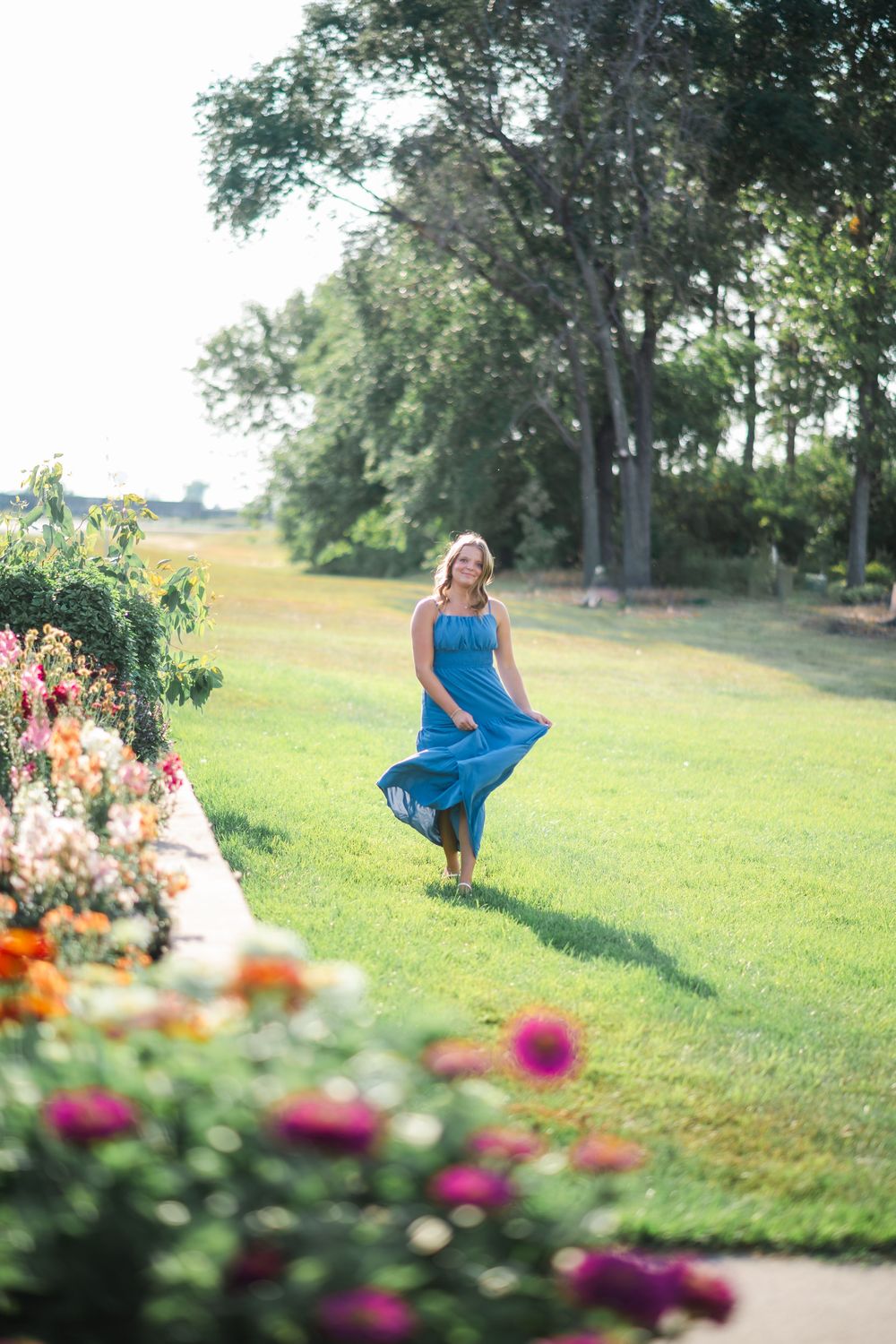 Woman in flowing teal dress twirling in lush green garden with colorful flowers on sunny day.