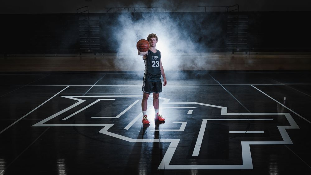 A basketball player dribbles on a dark court with dramatic lighting and white line markings creating geometric patterns.
