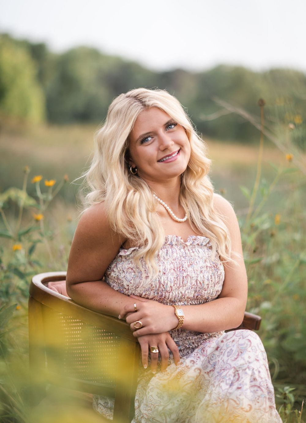 Young blonde woman in floral dress sitting in a wooden chair outdoors in a sunlit field with wildflowers.