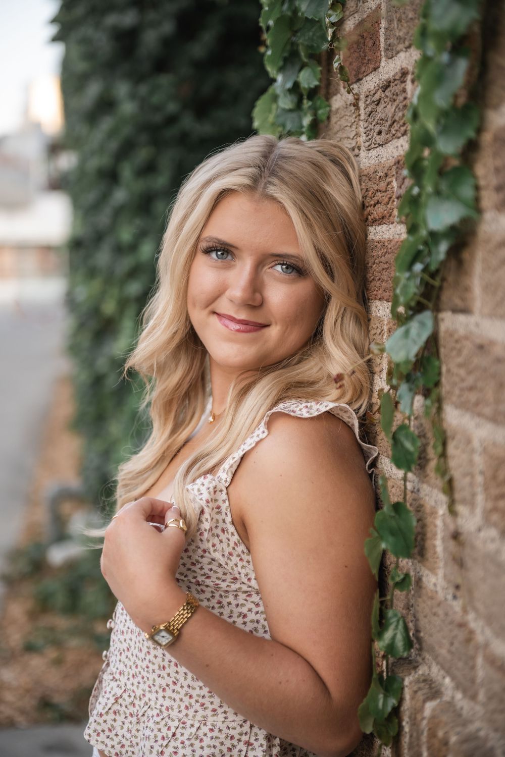 Young blonde woman in floral dress leaning against ivy-covered brick wall, smiling outdoors.