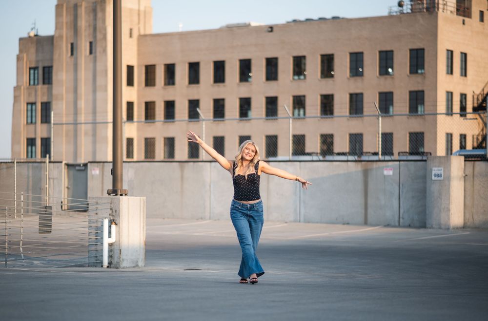 Woman in flare jeans and black tank top posing with arms outstretched on rooftop with urban building backdrop.