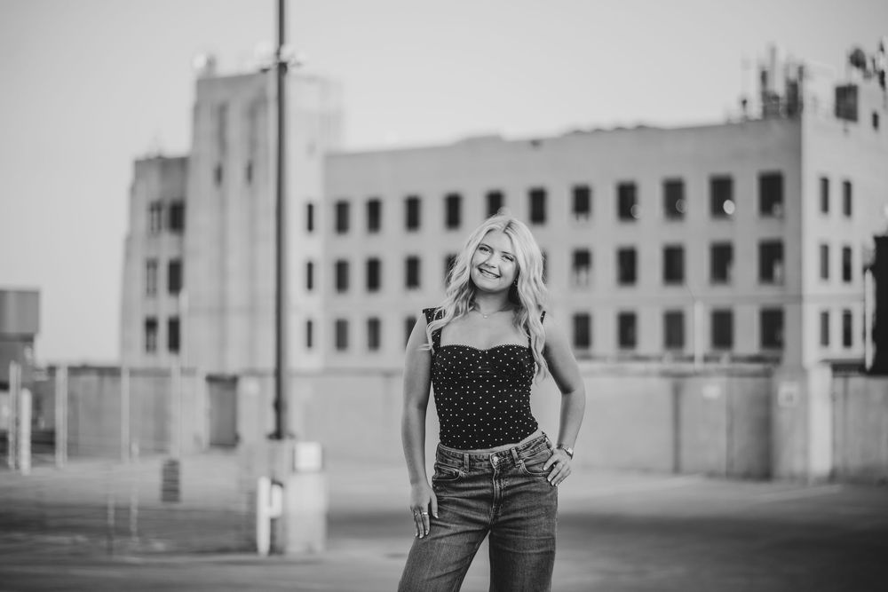 Smiling blonde woman in jeans and polka dot top poses in black and white photo with urban building backdrop.