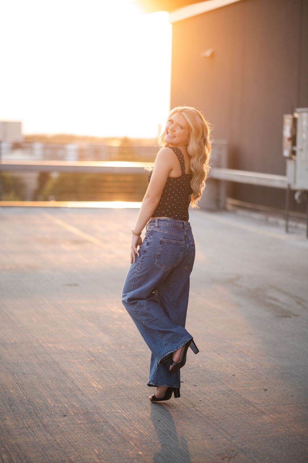Smiling blonde woman in wide-leg jeans and black top posing on rooftop during golden hour sunset.