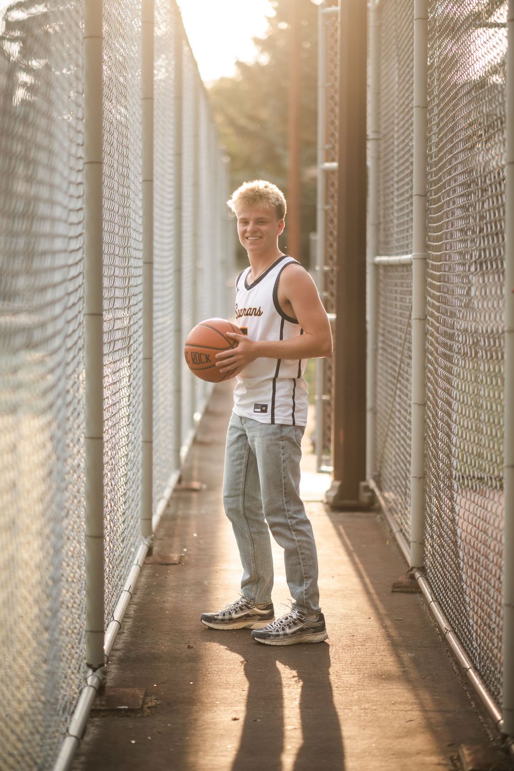 Smiling teen boy holding basketball in jersey and jeans between chain-link fences in golden hour sunlight.