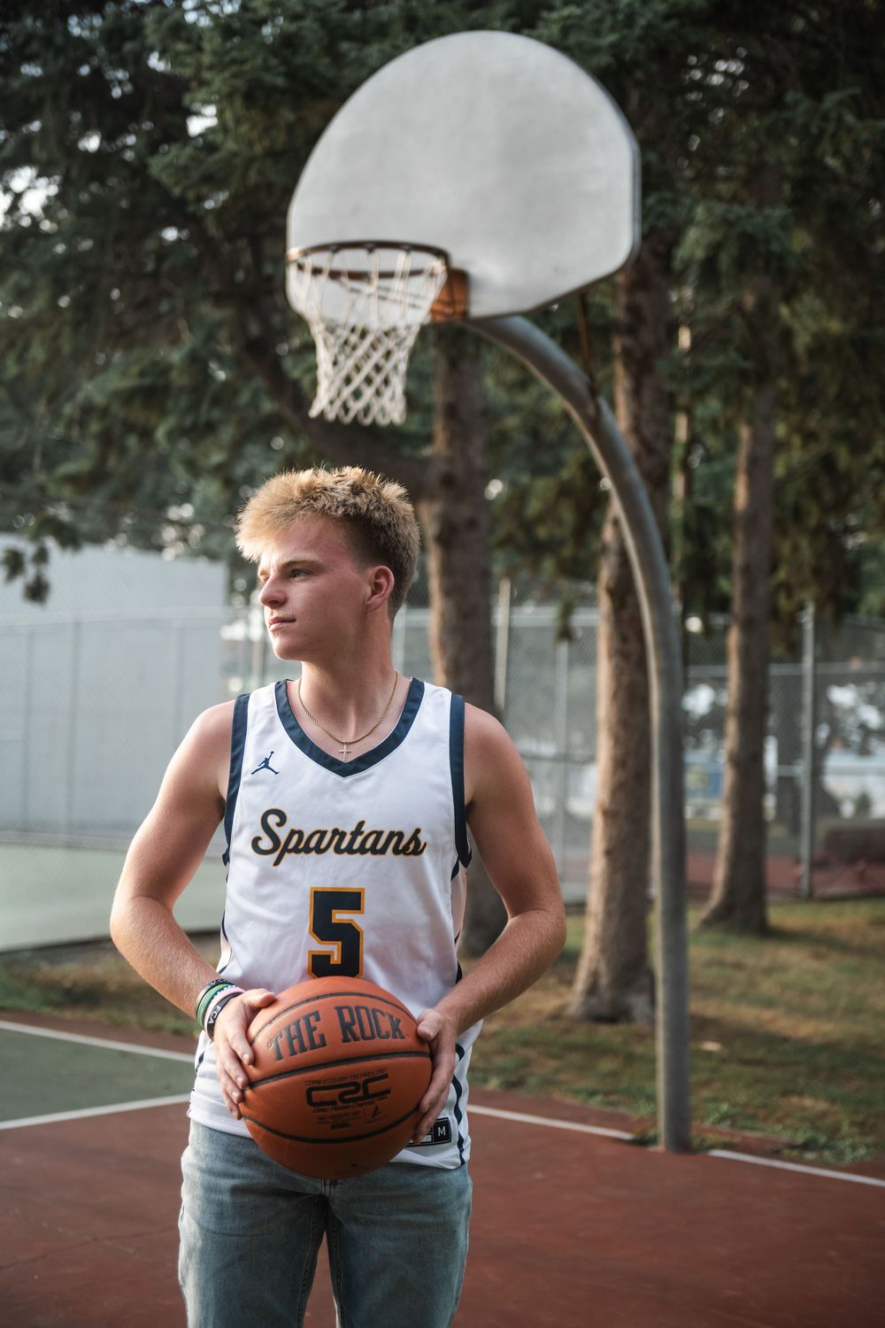 Young basketball player in Spartans #5 jersey holding a ball on an outdoor court with hoop in background.