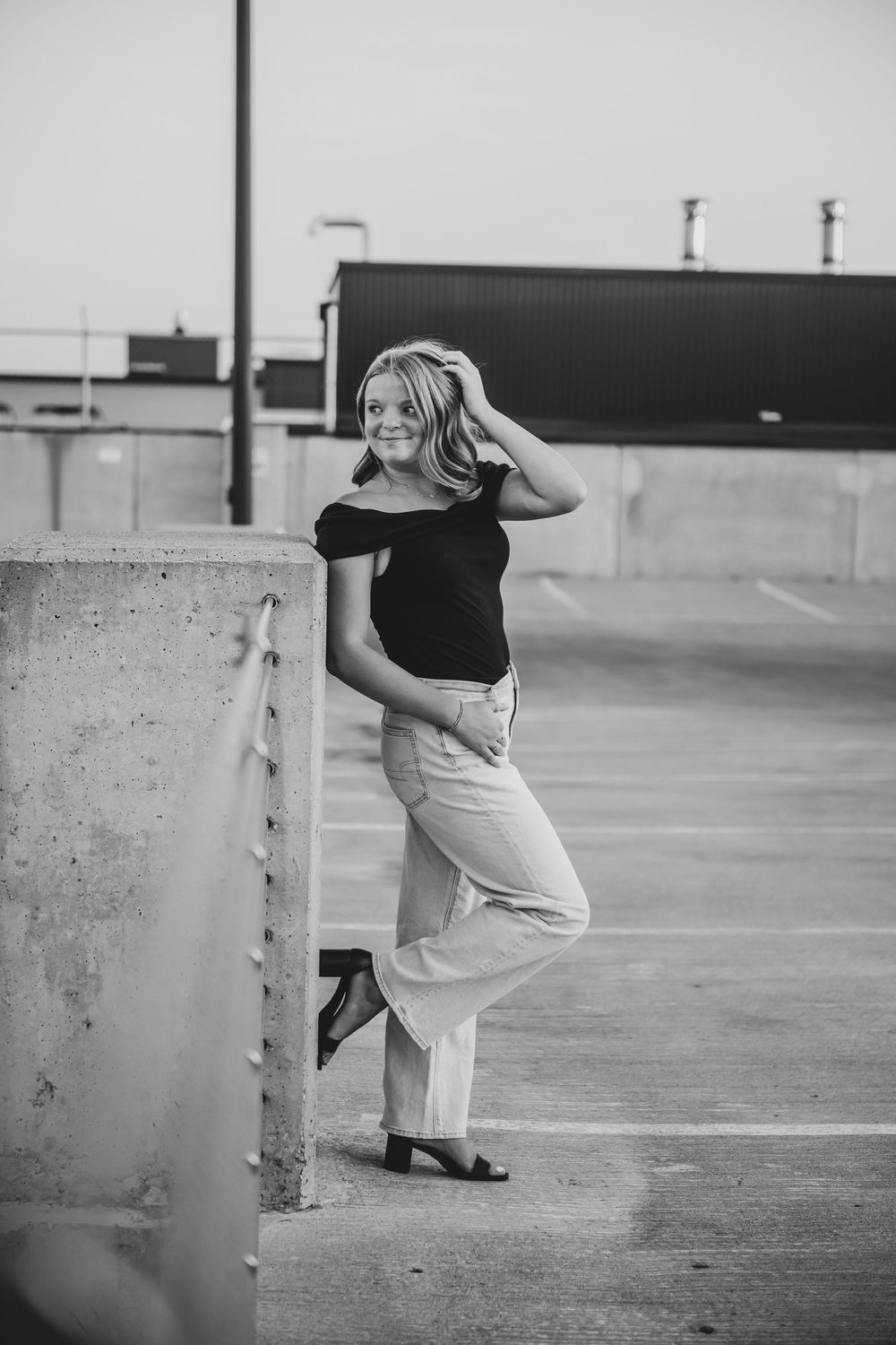 Black and white photo of a smiling young woman leaning against a concrete wall in a parking garage.