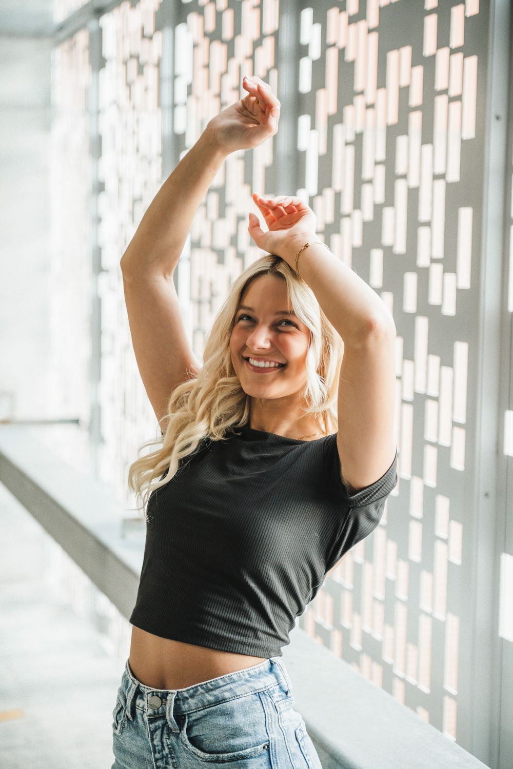 Joyful dance pose with arms raised against geometric white wall pattern.