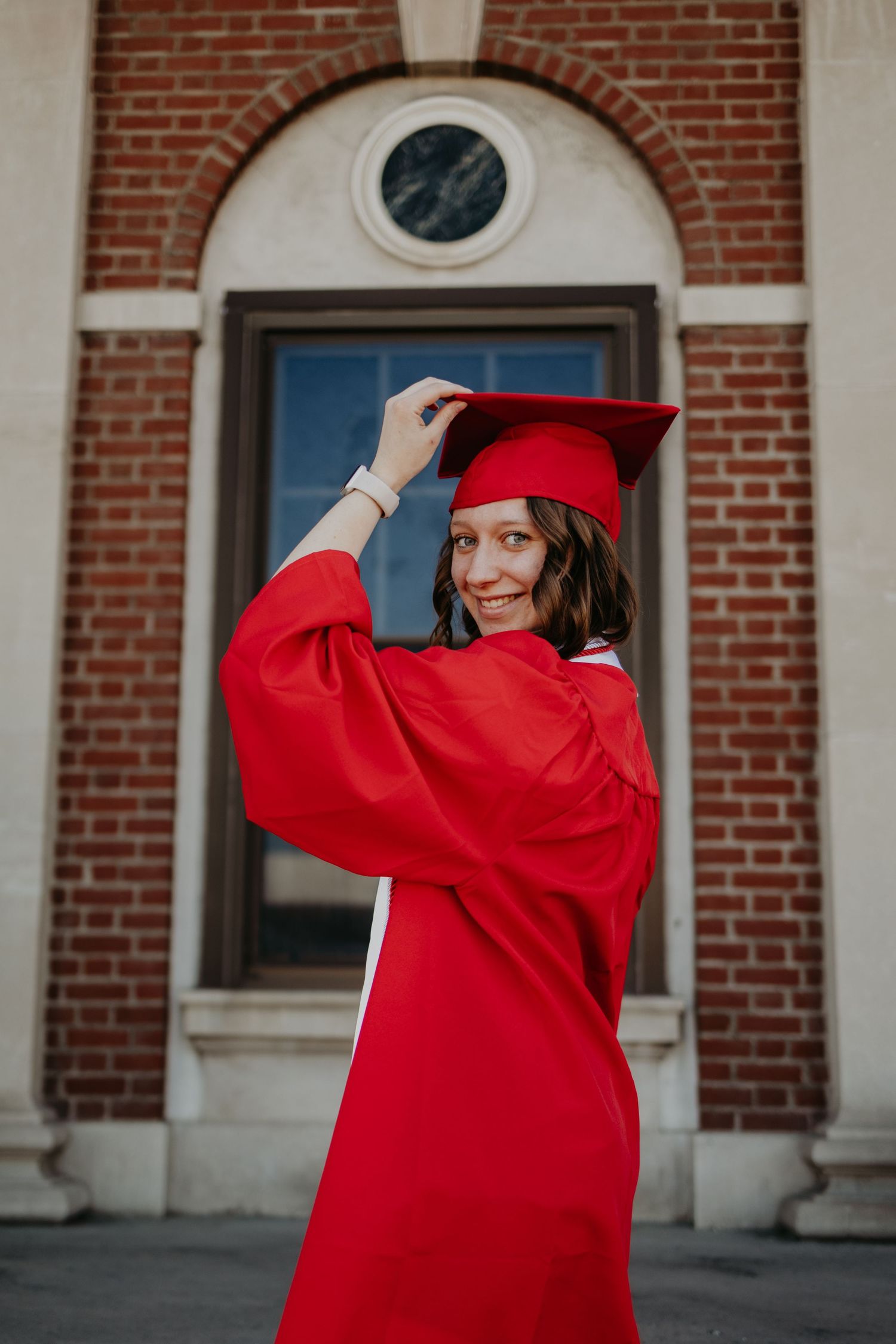 Graduate in red academic regalia playfully posing outside brick university building.