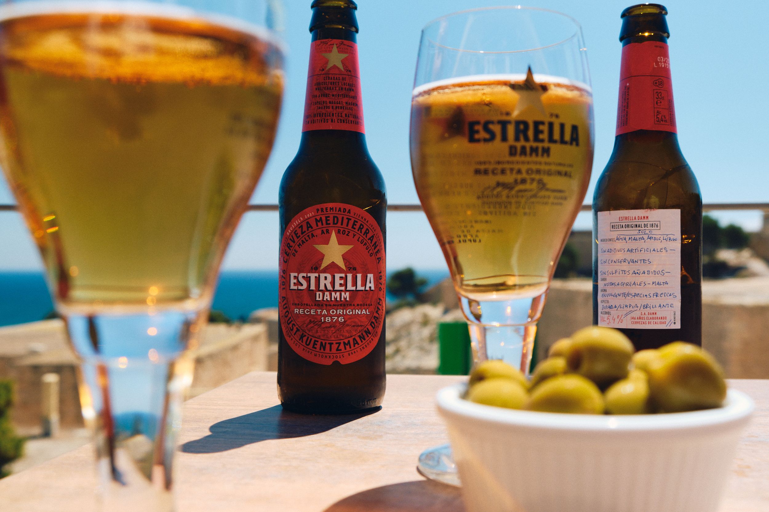 Cold glasses of Estrella beer served with olives on a sunny seaside terrace.