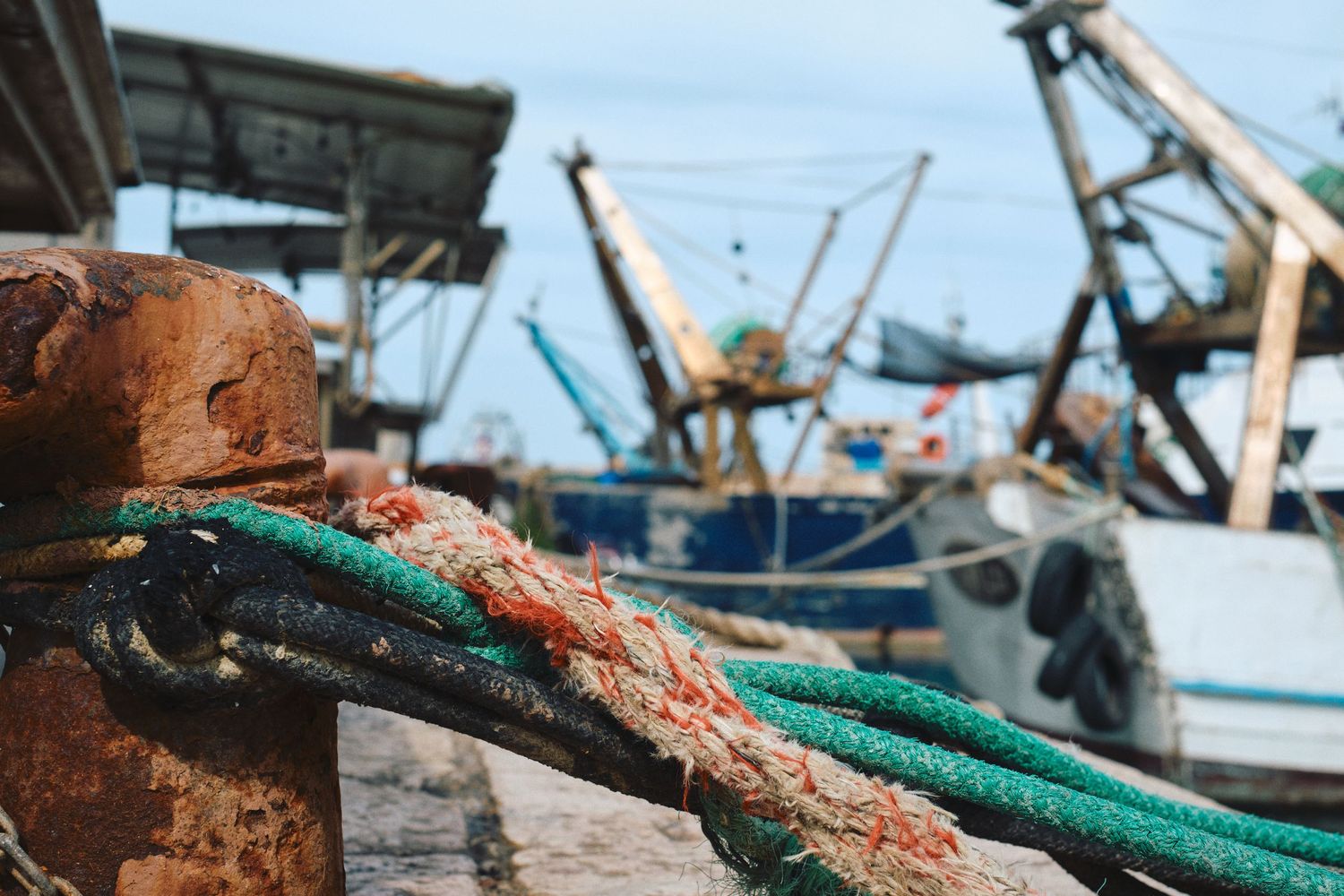 Rusty mooring hardware with green rope on a dock with fishing boats in background.