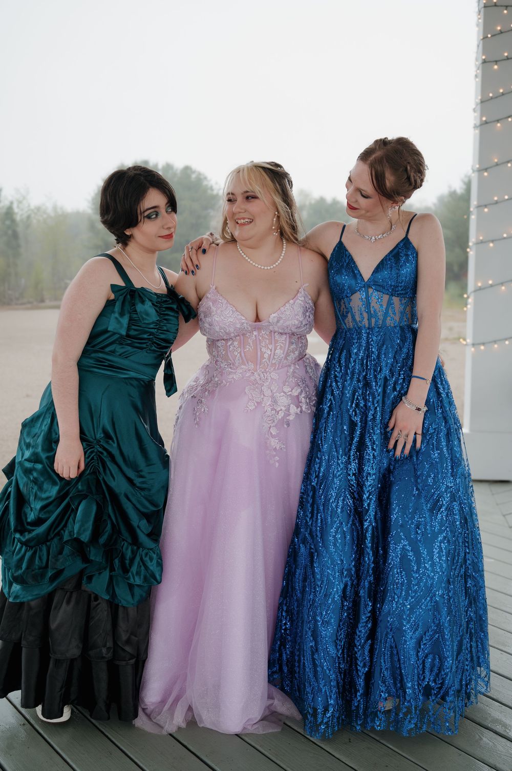 Three friends share intimate moment in formal dresses on outdoor deck.