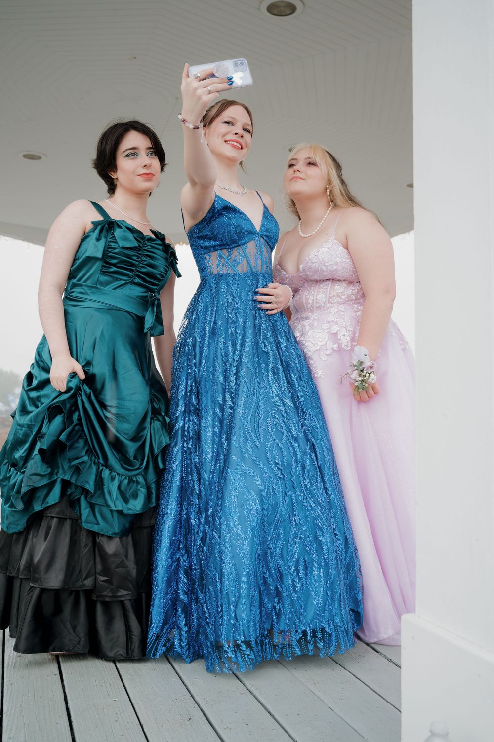 Three friends in formal dresses - pink, blue, and green - pose together for a photo on a porch.