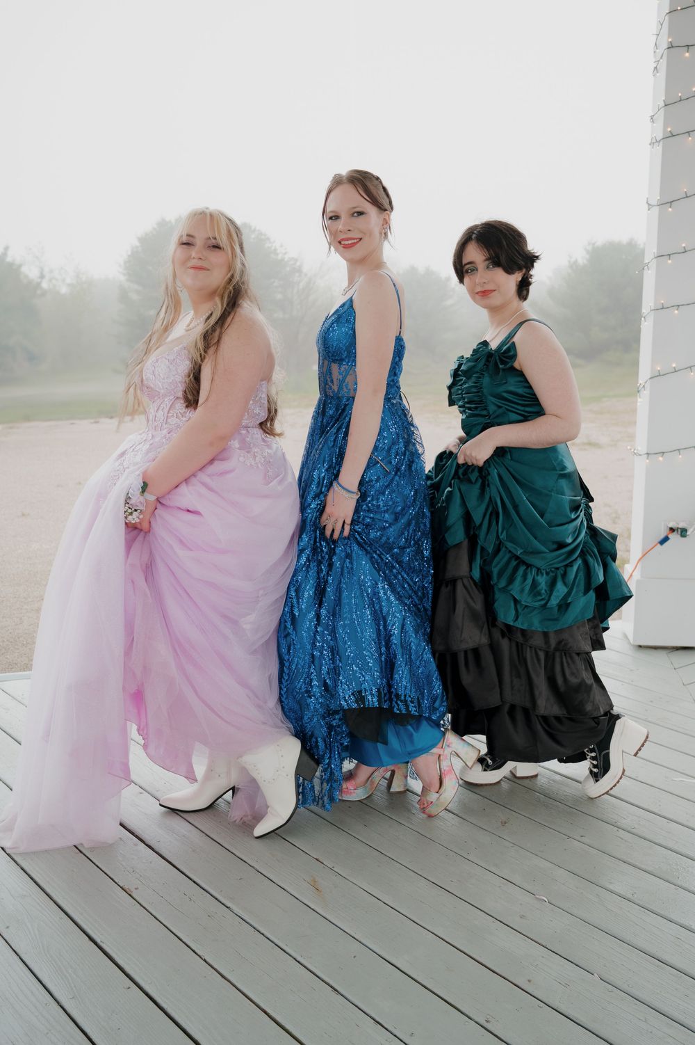 Three people in formal dresses pose together on a wooden deck in pink, blue, and emerald green gowns.