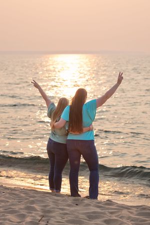 Three friends in matching blue shirts raise their arms in celebration while watching sunset at the beach.