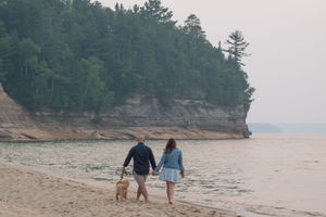 Two people walk their dog along a sandy beach with towering cliffs and evergreen trees in the background.