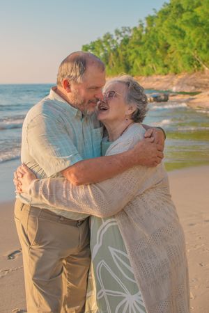 A tender embrace between two people on a sunny beach with water and trees visible in the background.