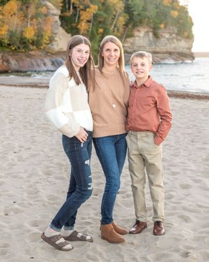 Three people in casual fall attire pose together on a sandy beach with cliffs and autumn foliage in the background.
