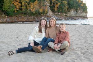 Three people sitting casually together on a sandy beach with cliffs and autumn foliage in the background.
