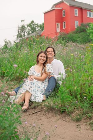A couple sits together in a wildflower meadow with a red house in the background during a summer photoshoot.