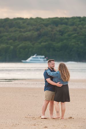 A couple embraces on a sandy beach at sunset with a boat visible in the background on the water.