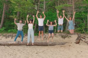 A group of people jumping joyfully on a sandy beach with a green forest backdrop in summer.