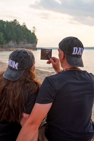 Two people in matching black caps sit side by side watching the sunset over a lake.