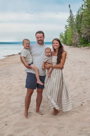 A family poses together on a sandy beach with pine trees and water in the background.