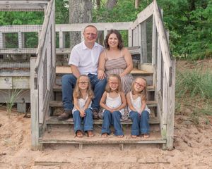 Family of five sitting together on wooden beach stairs surrounded by sand and greenery.