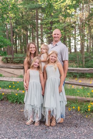 A family wearing coordinated white and gray outfits poses together on a gravel path surrounded by trees and flowers.