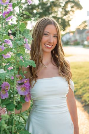 A figure in a white dress poses among pink hollyhock flowers during golden hour in a garden setting.
