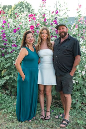 Three people pose together in a garden with blooming pink hollyhock flowers creating a beautiful summer backdrop.